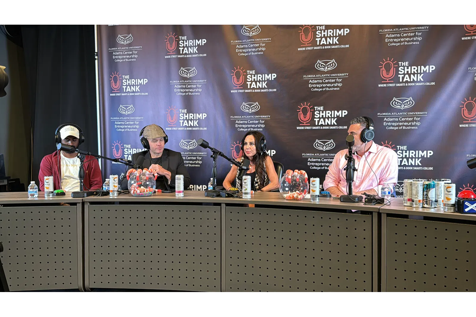 Four hosts wearing headphones speak into microphones during The Shrimp Tank podcast recording with branded backdrop.