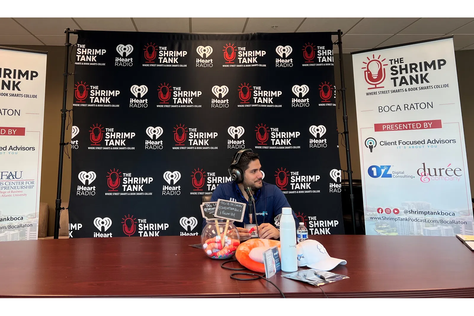 a person sitting at a table with a microphone and a sign behind him