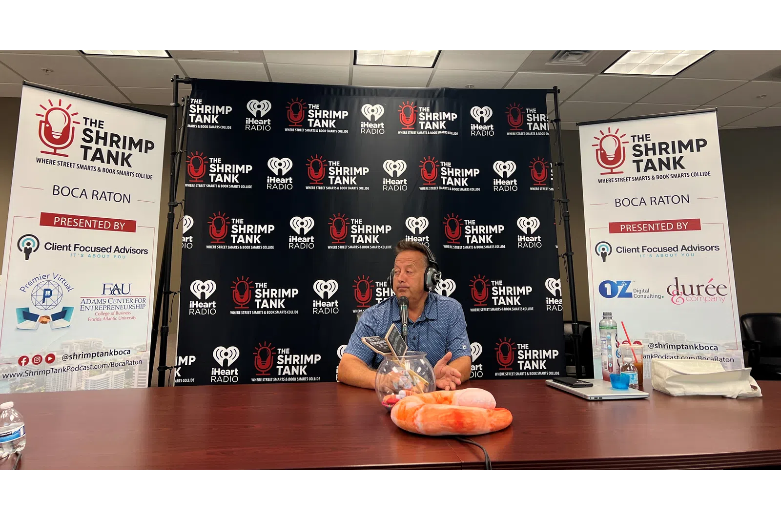 a person sitting at a table with a microphone and a sign behind him