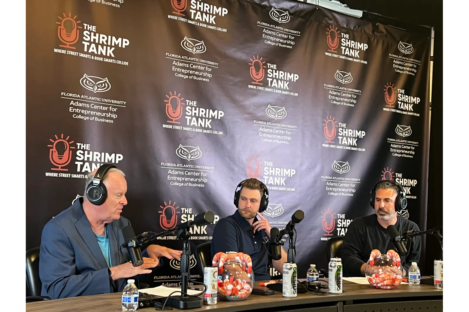 a group of men sitting at a table with microphones and a blackboard behind them