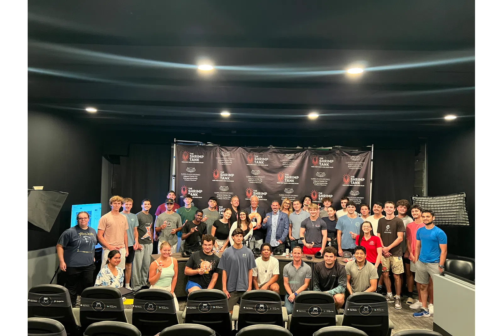 Large group of diverse young adults posing in front of Shrimp Tank banner in well-lit indoor event space.