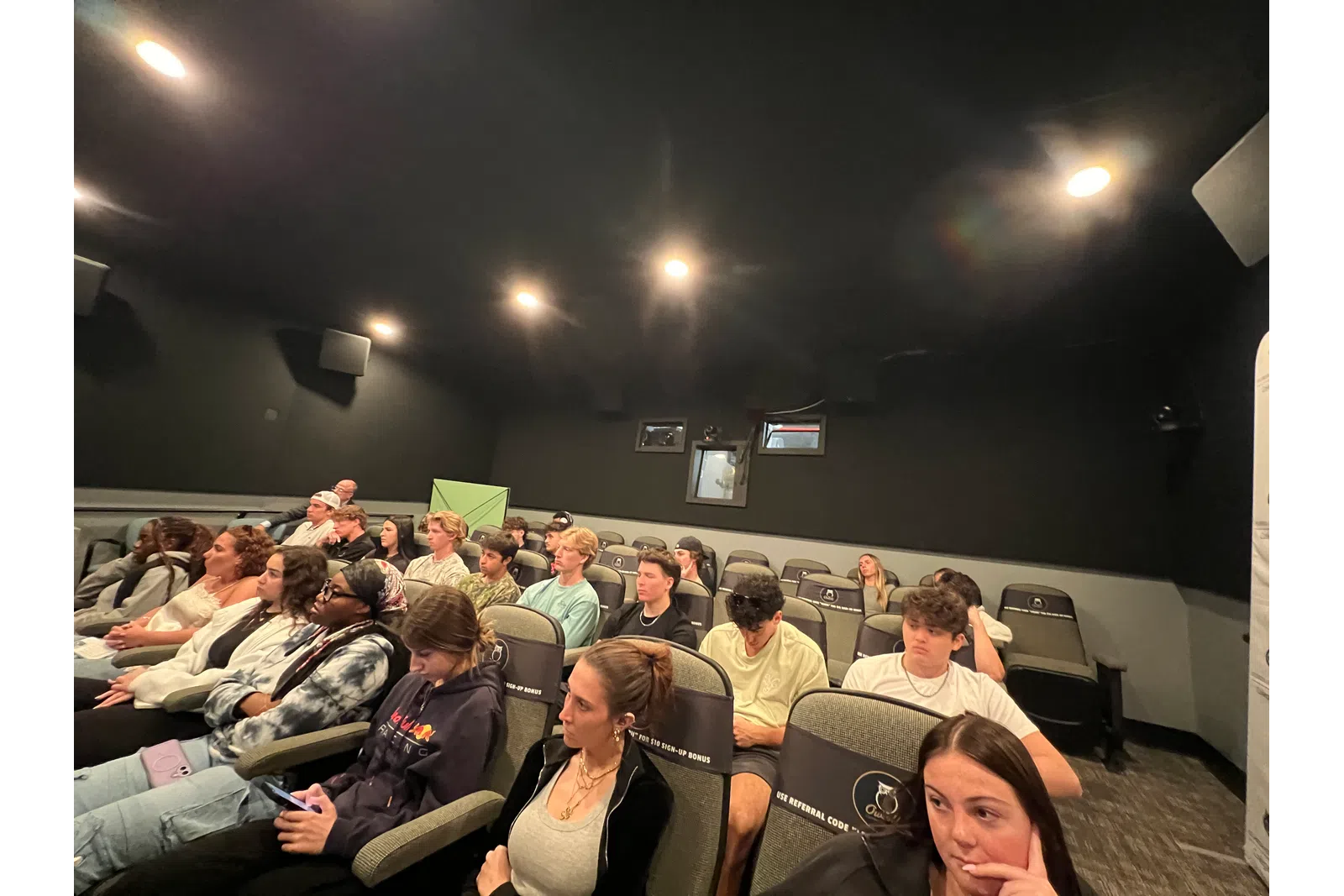 A diverse group of young adults attentively watching a presentation in a dimly lit auditorium with black walls.