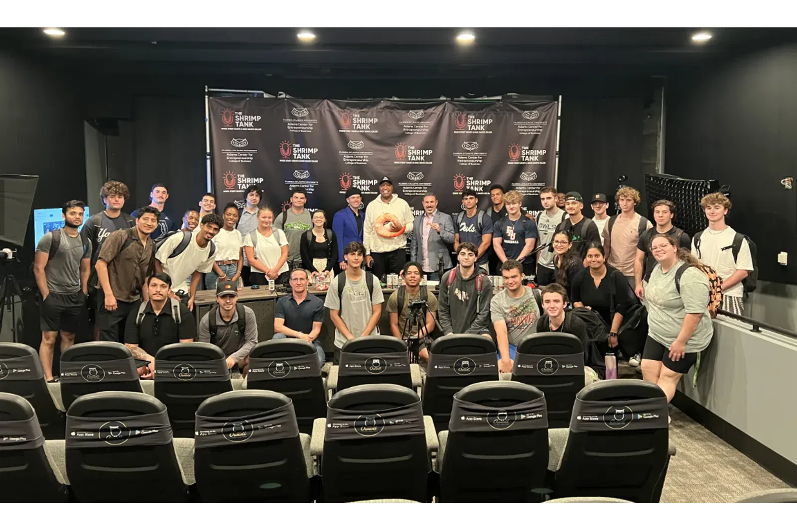 Group of young adults and instructors posing for a photo in a studio with Shrimp Tank logos on the background.