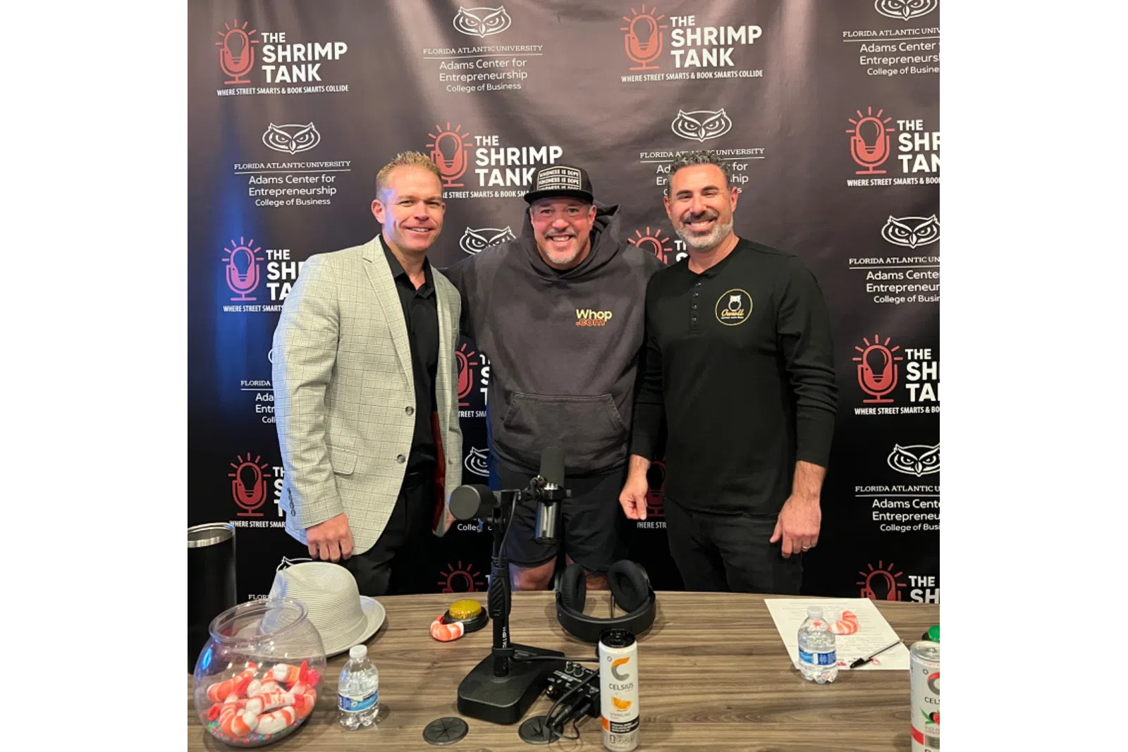 Three men posing in front of The Shrimp Tank podcast backdrop at a recording table with microphones and refreshments.