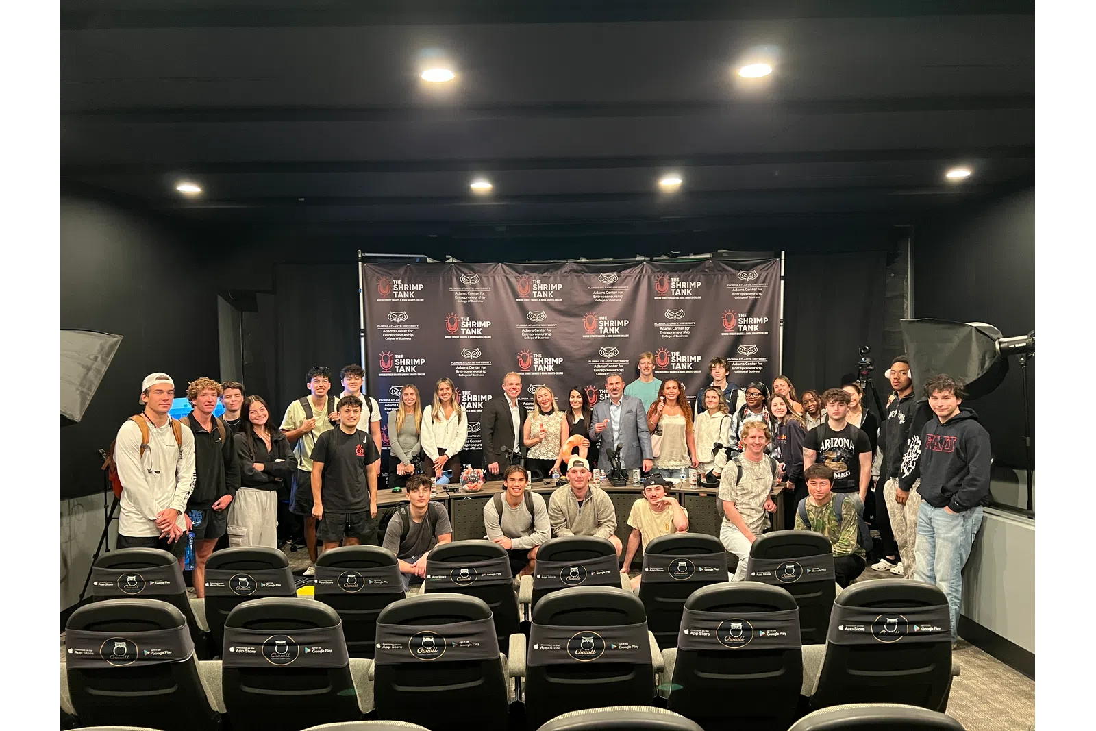 Group photo of diverse young people and adults posing in front of a branded backdrop in a studio setting.