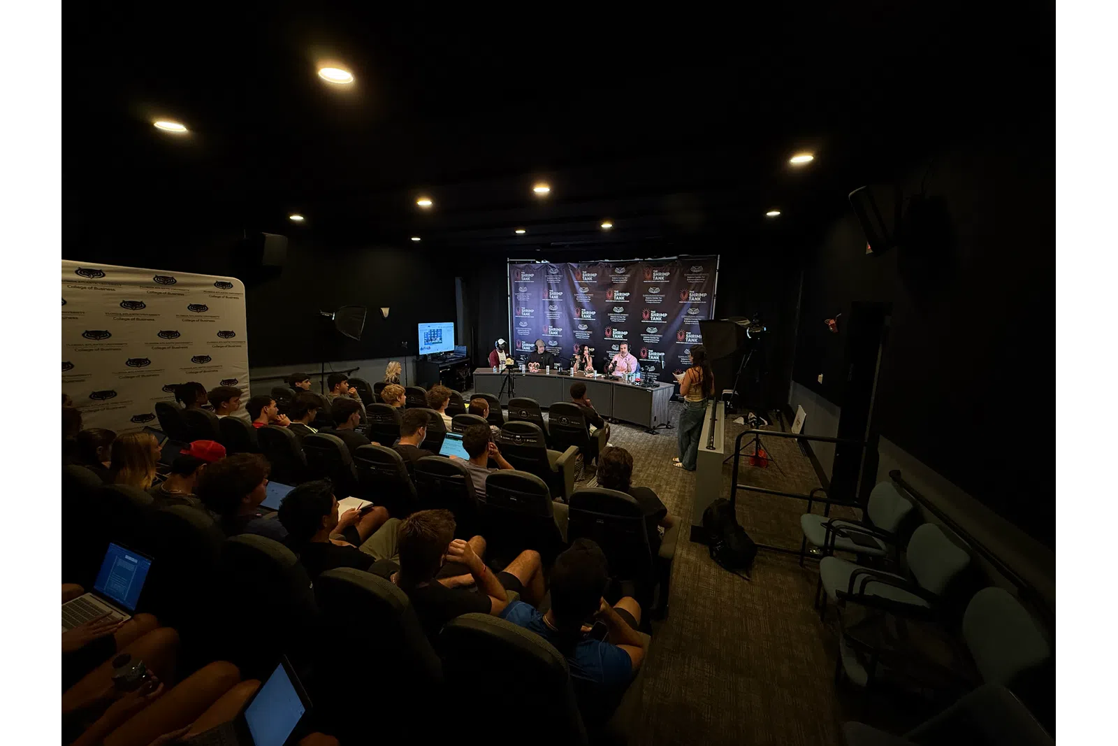 Audience attentively watching a panel discussion in a dark conference room with speakers at a table and media equipment.