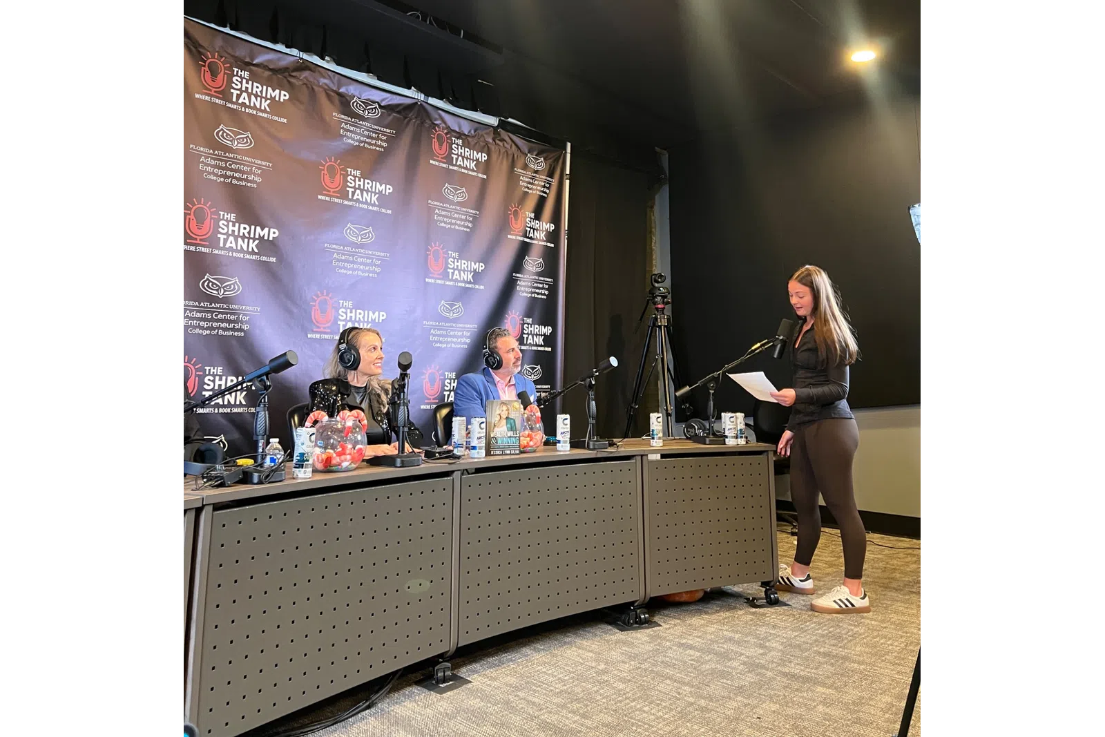 Radio hosts in headphones recording a podcast with a standing woman speaking into a microphone in a studio.