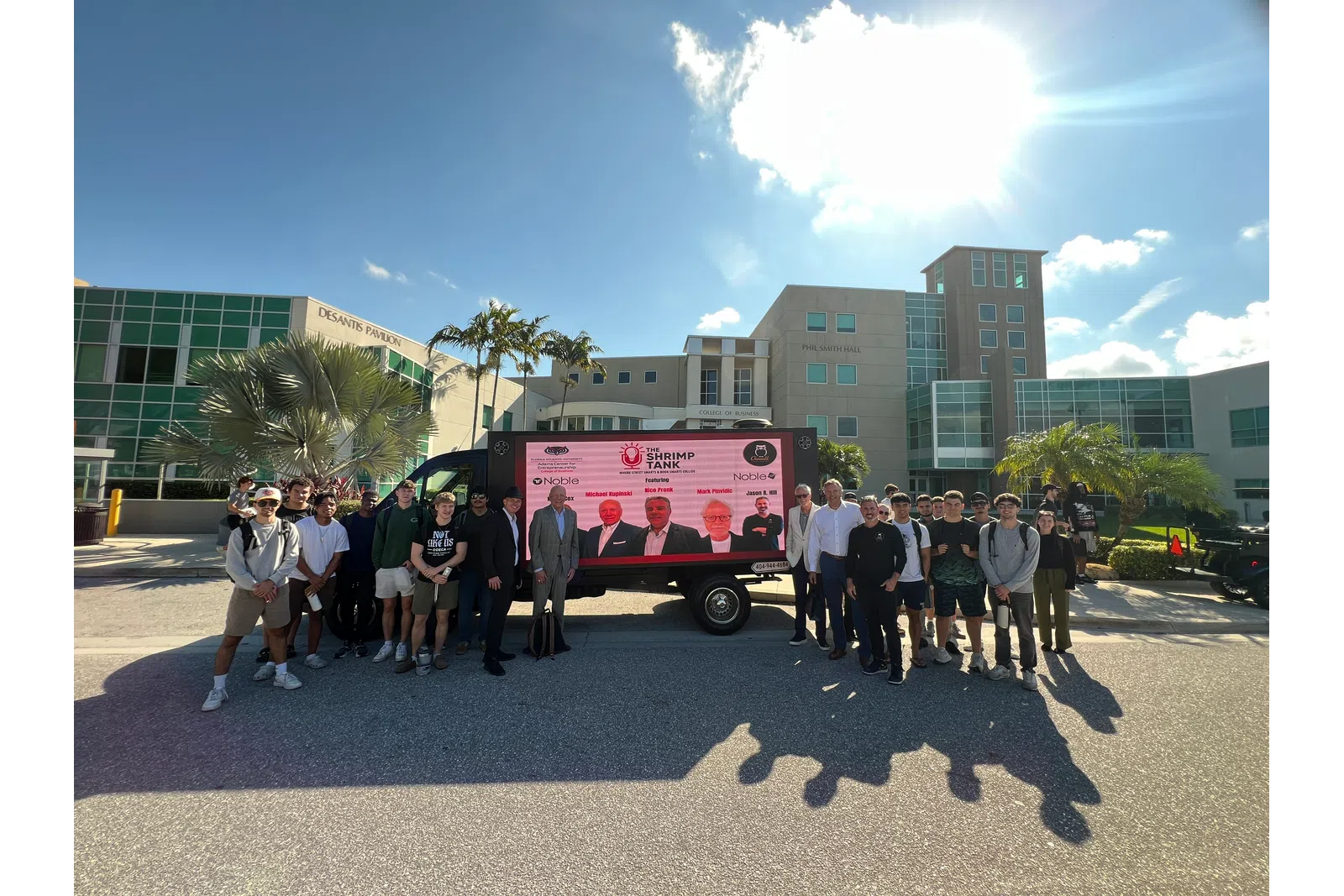 Group of people posing outdoors in front of a trailer with Shrimp Tank display at a sunny campus with palm trees.