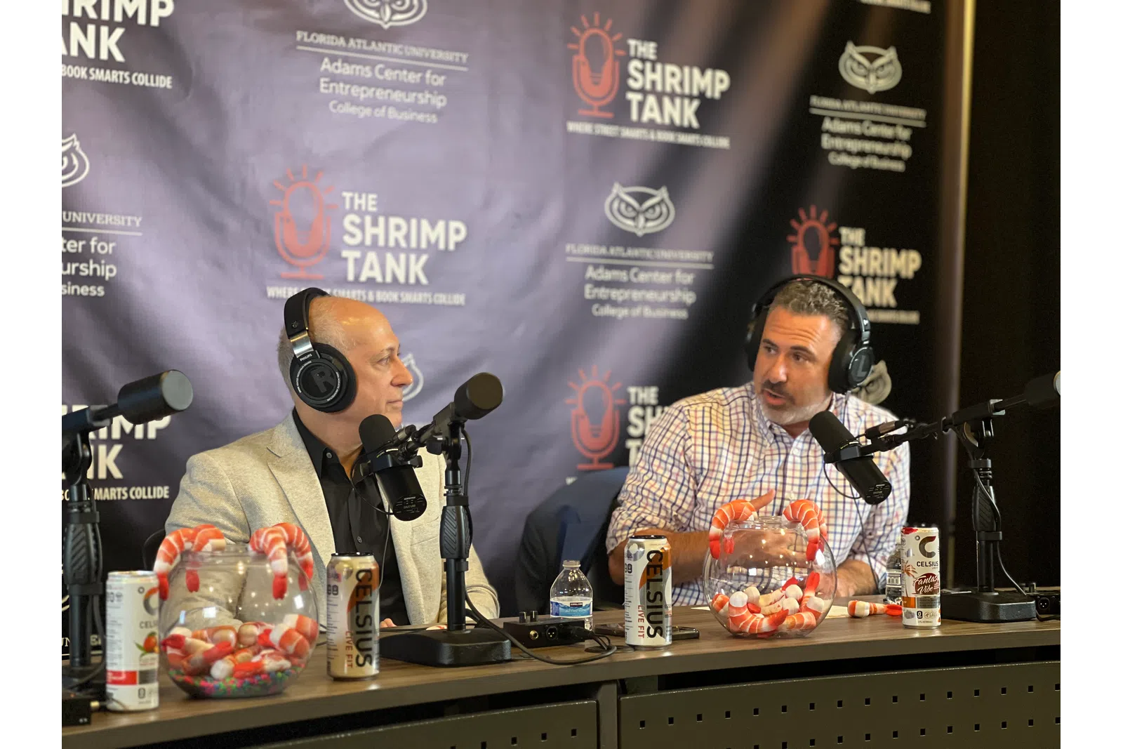 Two men wearing headphones discussing on a podcast set with shrimp and Celsius drink cans on the table