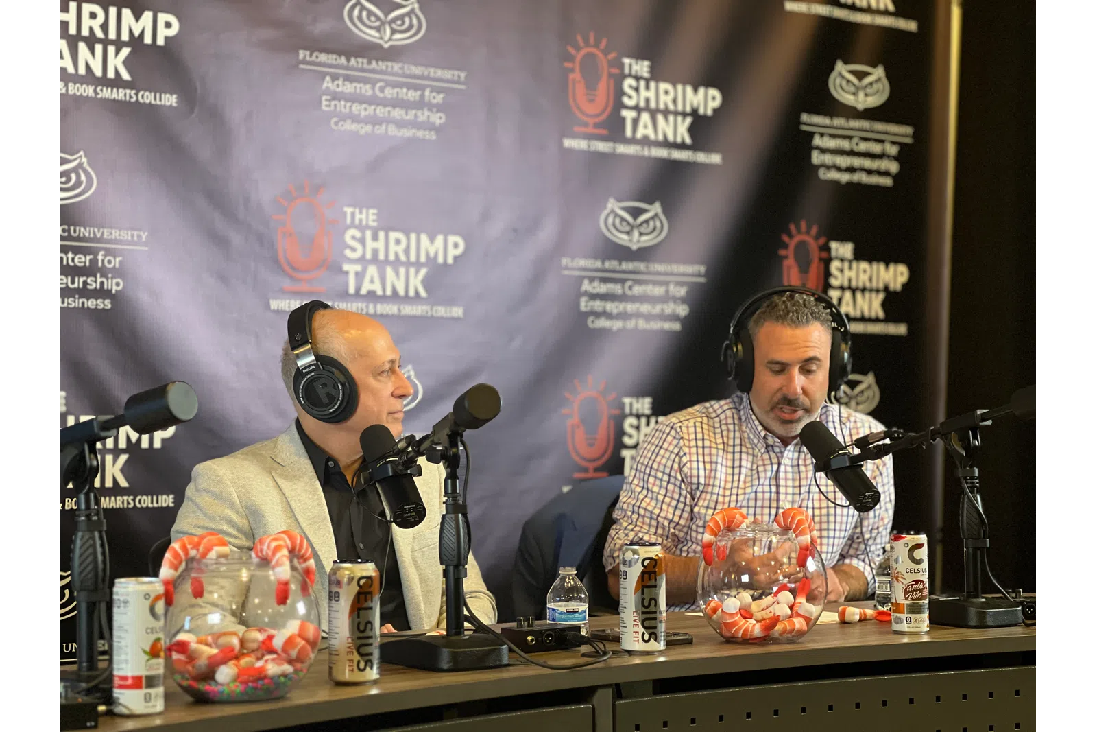 Two men with headphones speaking into microphones at The Shrimp Tank podcast with shrimp and soda on the table