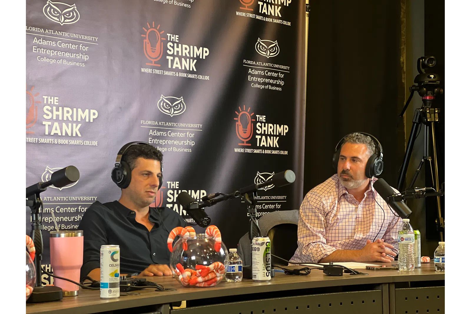 Two men podcasting on The Shrimp Tank with microphones, headphones, and branded backdrop in a studio setting.
