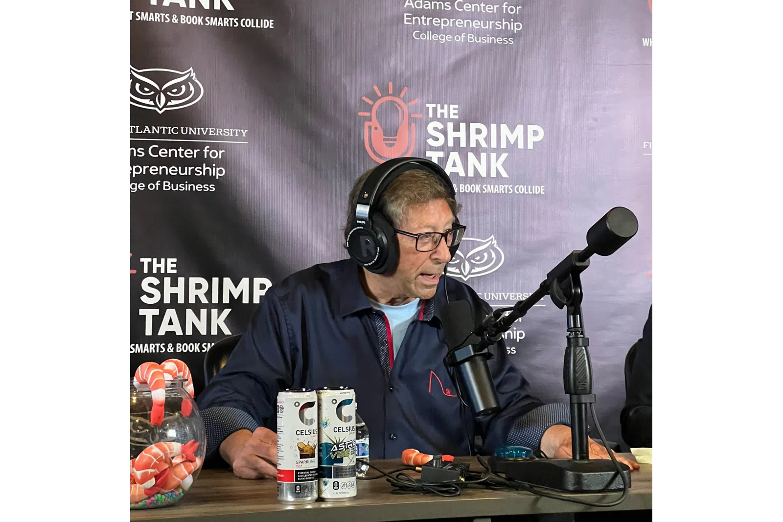 Man wearing headphones speaking into a microphone at The Shrimp Tank podcast with drink cans on the table