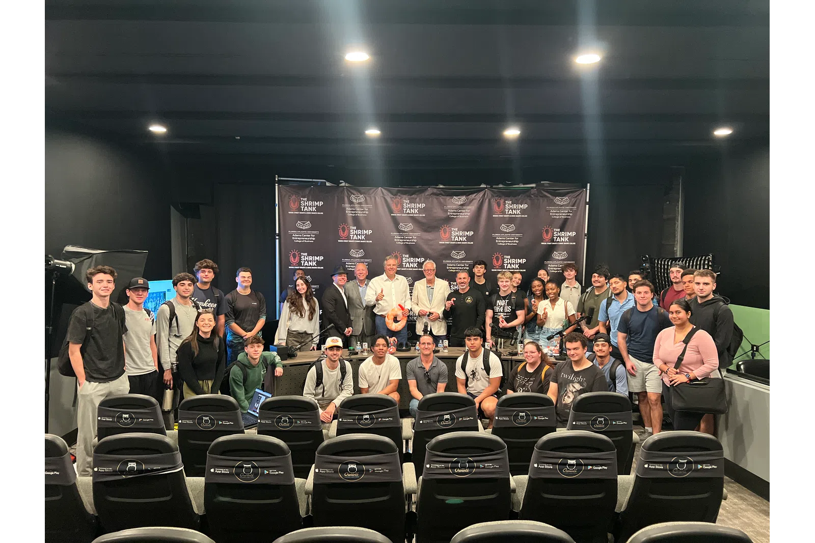 Large group of diverse young adults and speakers posing in front of a Shrimp Tank branded backdrop in auditorium.