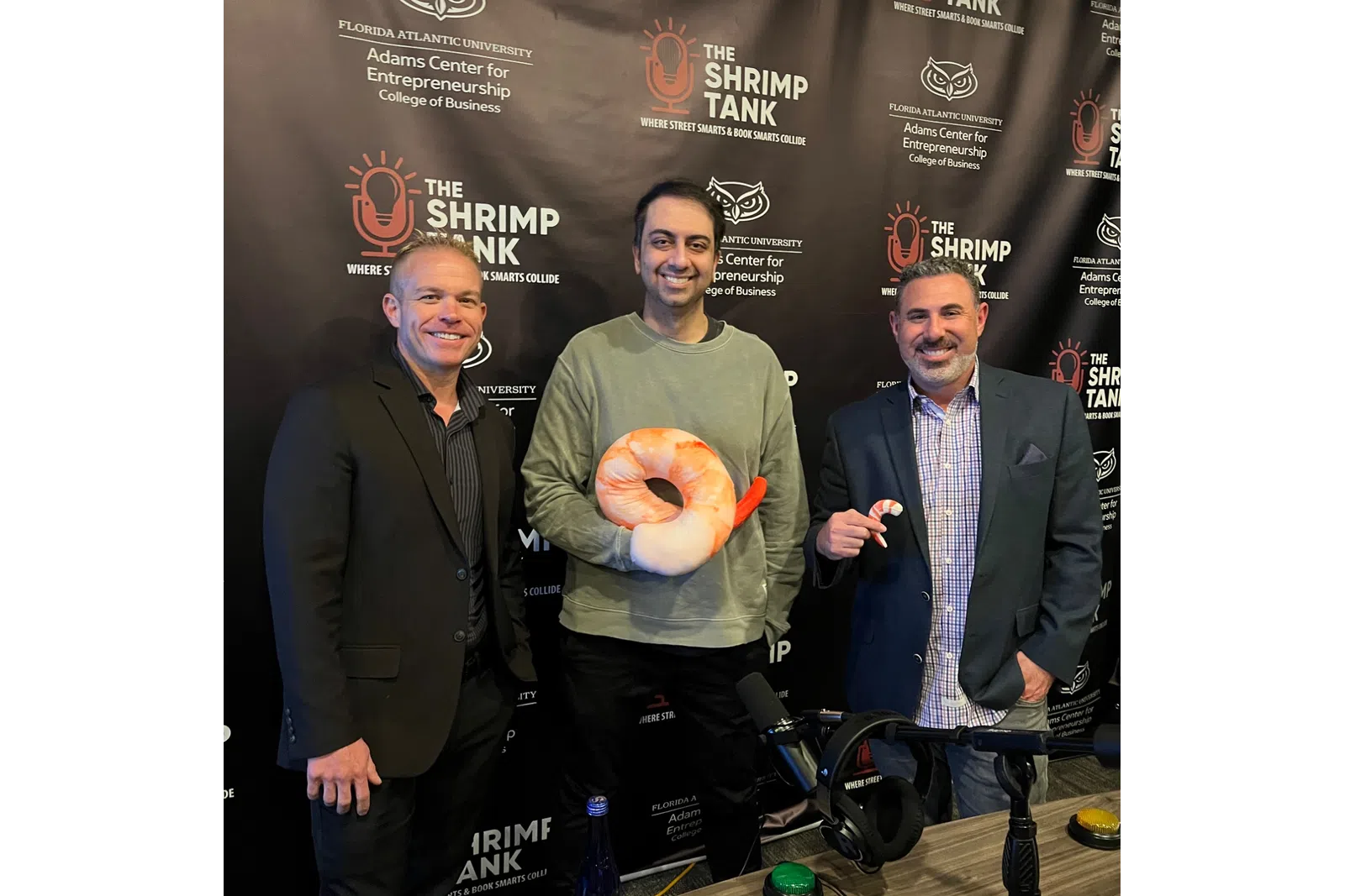 Three men posing at The Shrimp Tank event with shrimp plush toys and a branded backdrop of Florida Atlantic University.