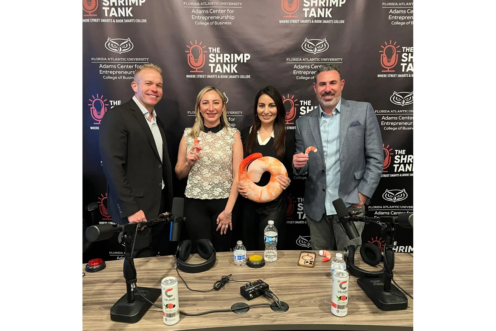Four people posing with shrimp-themed items at a podcast studio with The Shrimp Tank backdrop.