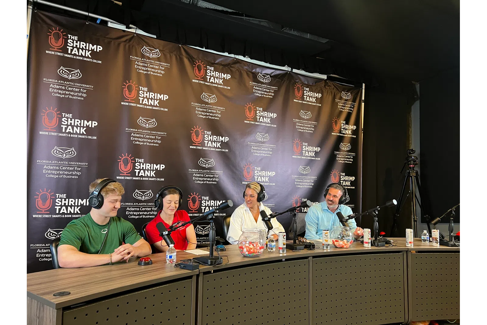 Four hosts with microphones and headphones recording podcast at The Shrimp Tank studio with branded backdrop.