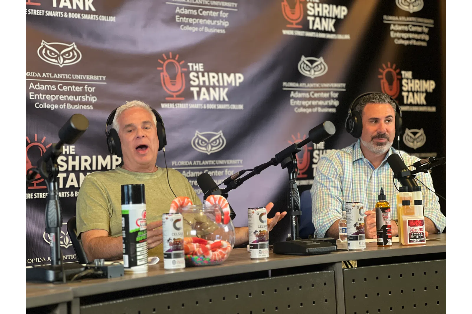 a couple of men sitting at a table with microphones and food