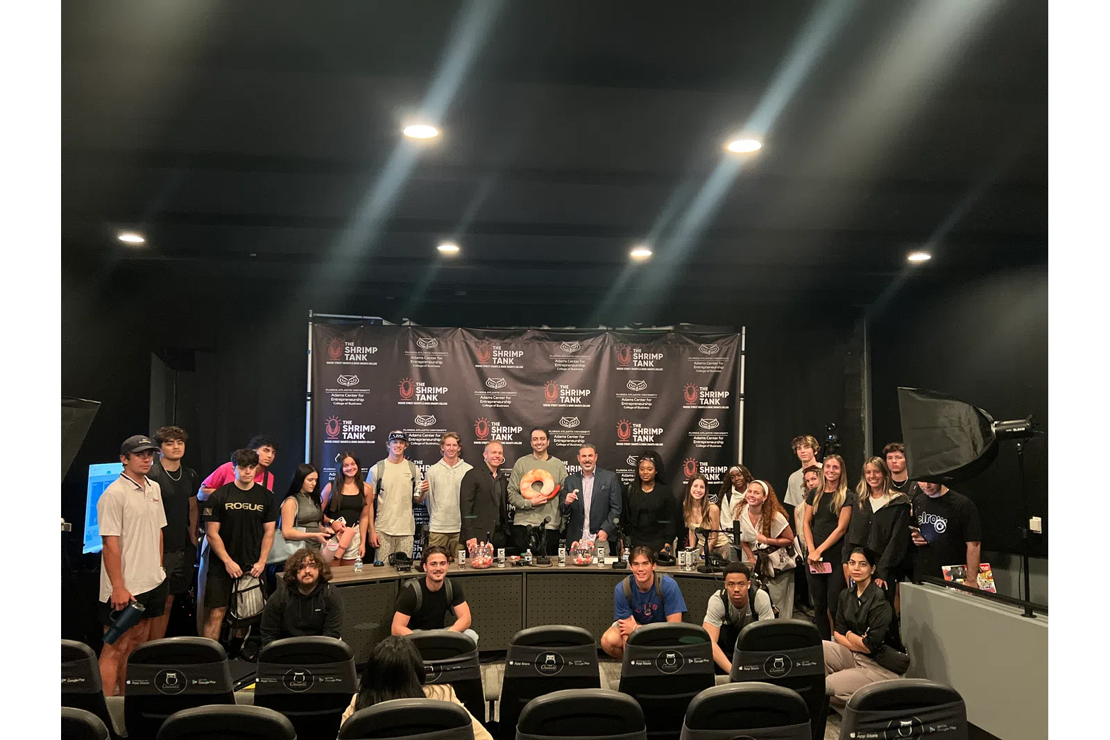 Group of people posing in a studio with Shrimp Tank banners and professional lighting equipment in the background.