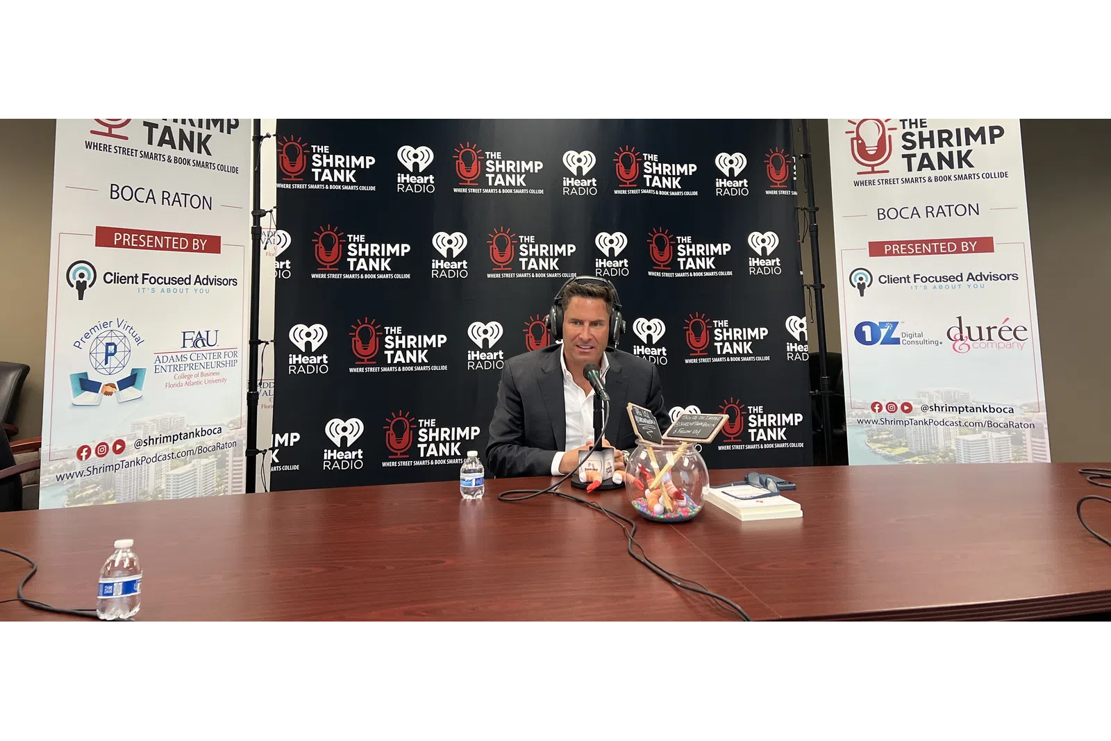 a person sitting at a table with microphones and a sign behind him