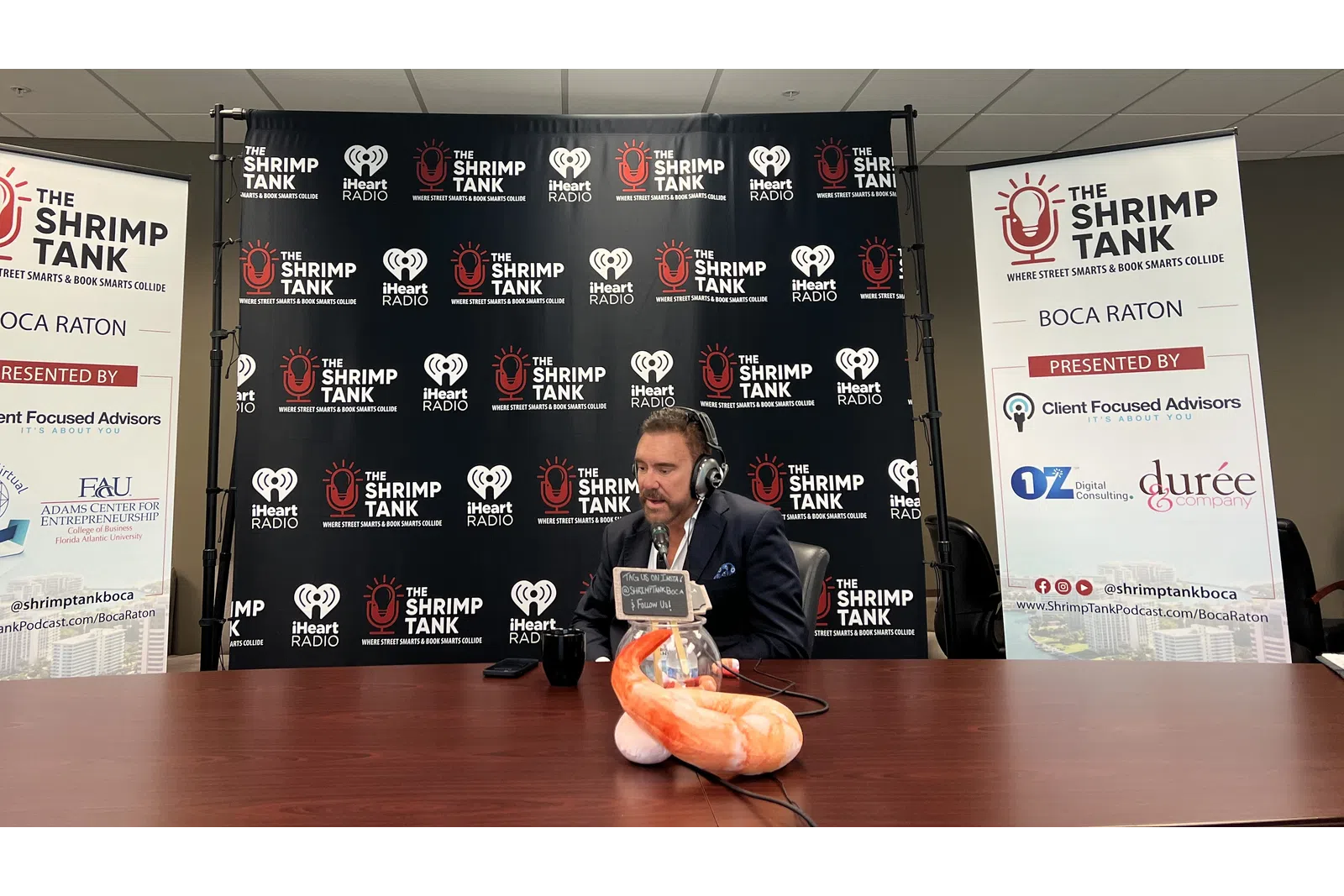 a person sitting at a table with a microphone and a sign behind him