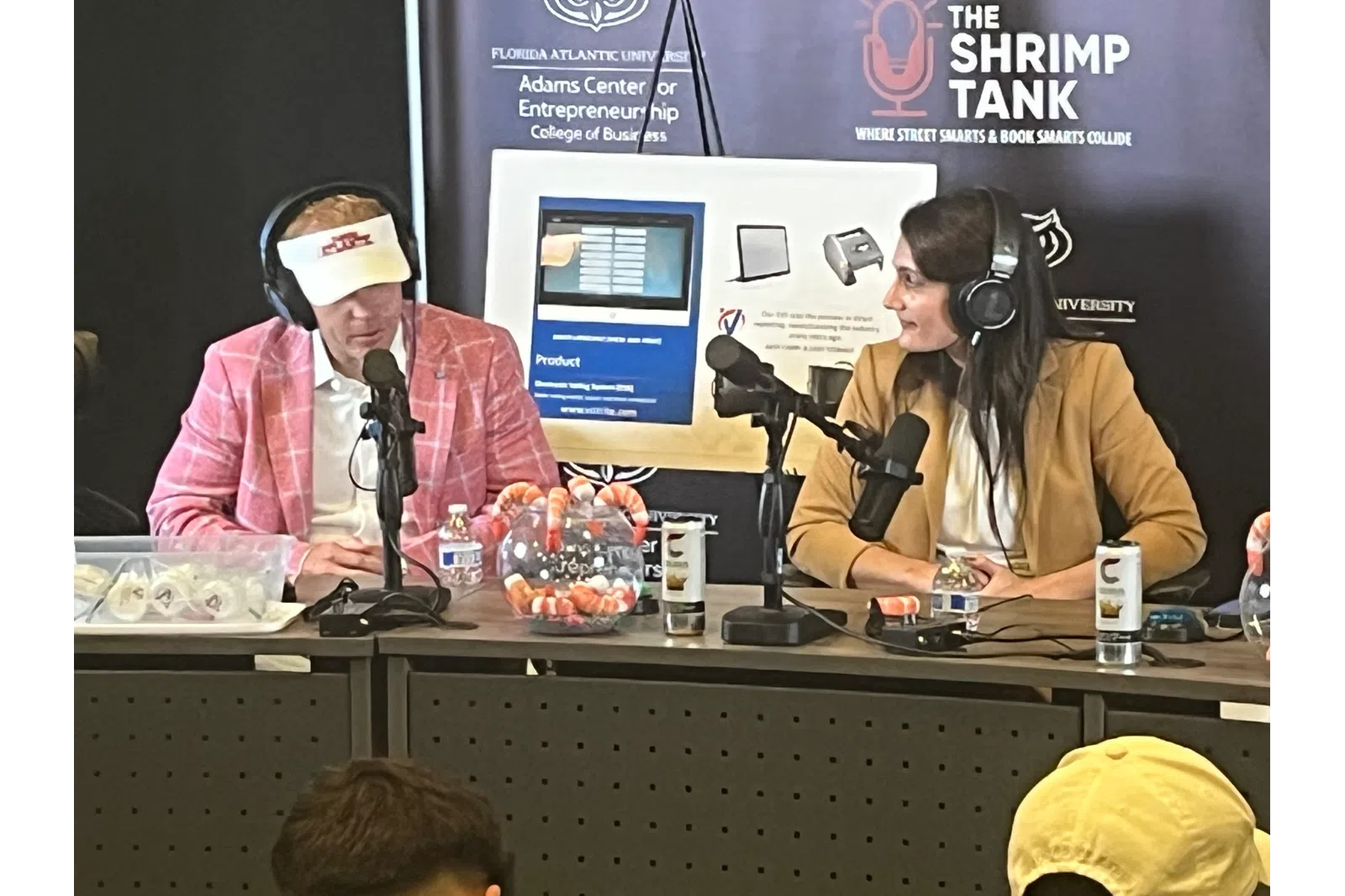 Two podcast hosts wearing headphones converse at a table with microphones and shrimp decor, Shrimp Tank event backdrop.