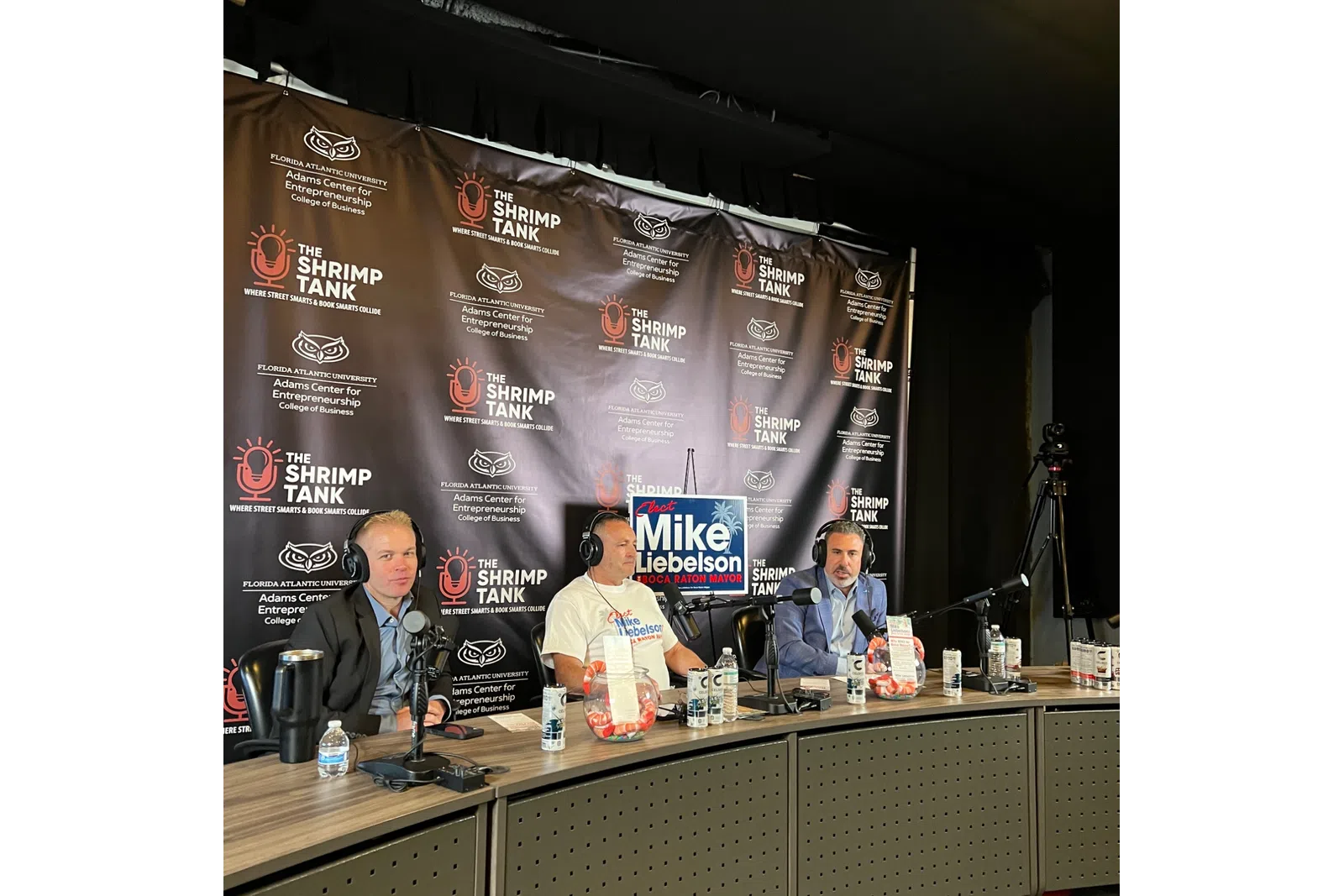 Three men recording a podcast at The Shrimp Tank with microphones, headphones, and a campaign sign for Mike Rodelson.