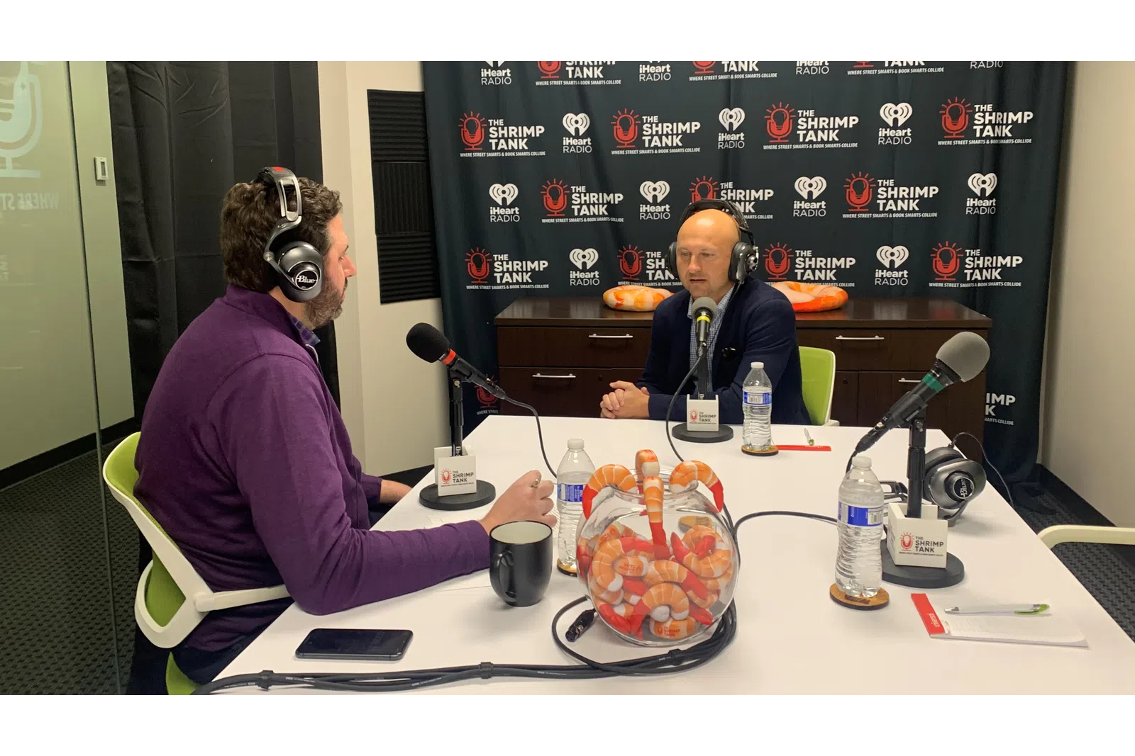 a person wearing headphones sitting at a table with a microphone and a person behind him