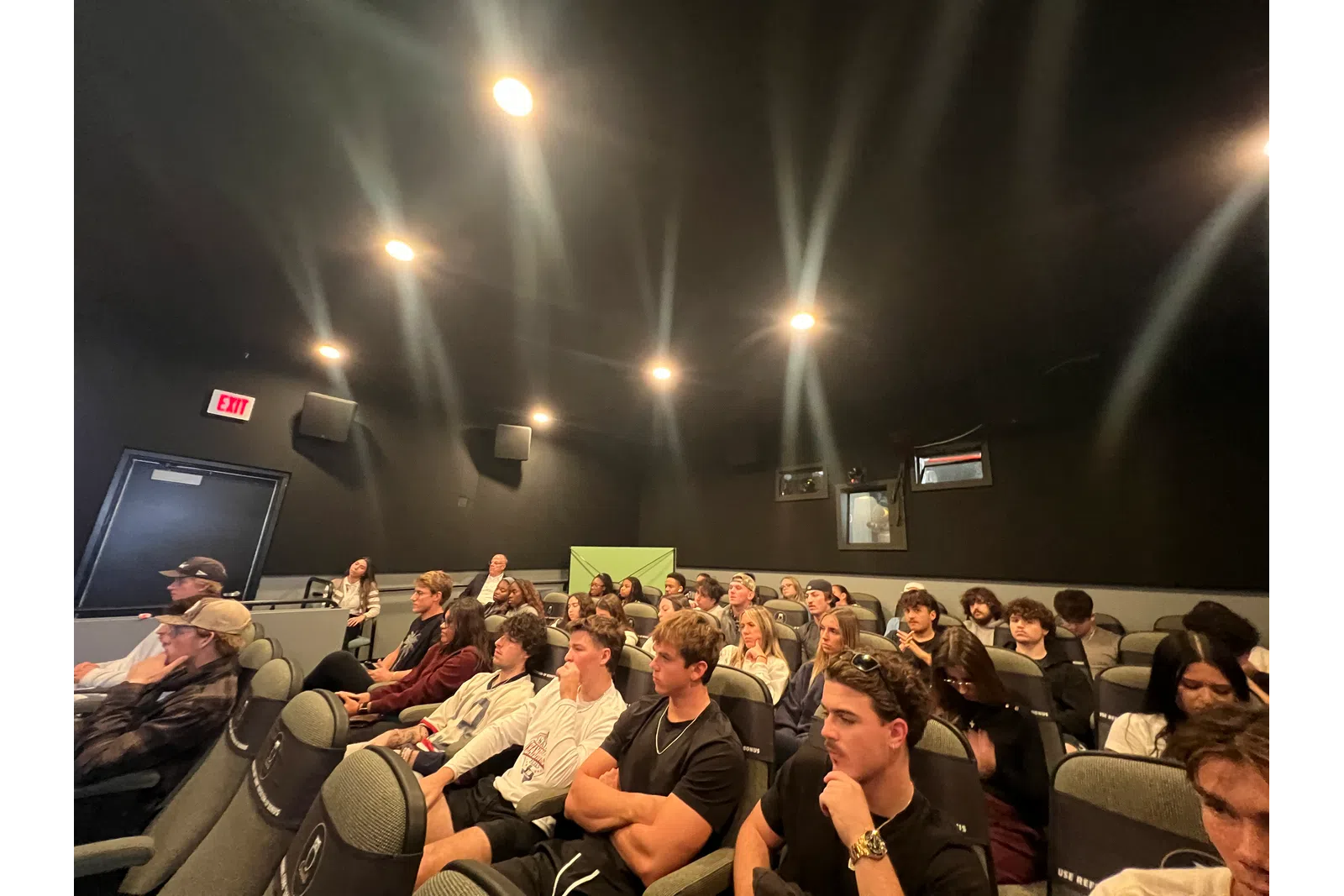 Audience seated and attentively watching in a small, dark theater with overhead spotlights.