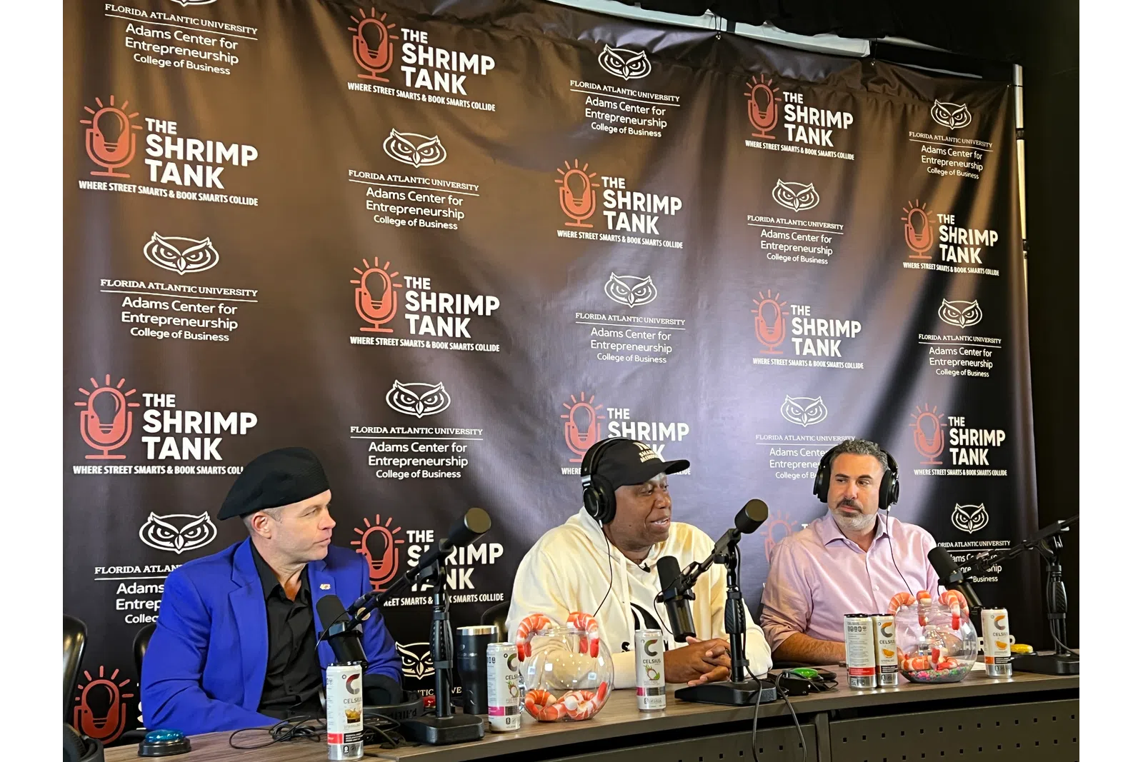 Three men speaking into microphones during The Shrimp Tank podcast with branded backdrop and shrimp bowls.