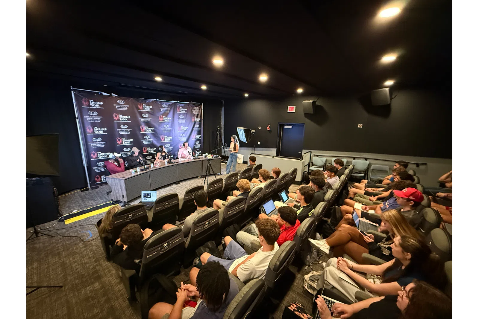 Conference room with panel discussion, attendees in theater seats using laptops and cameras recording event