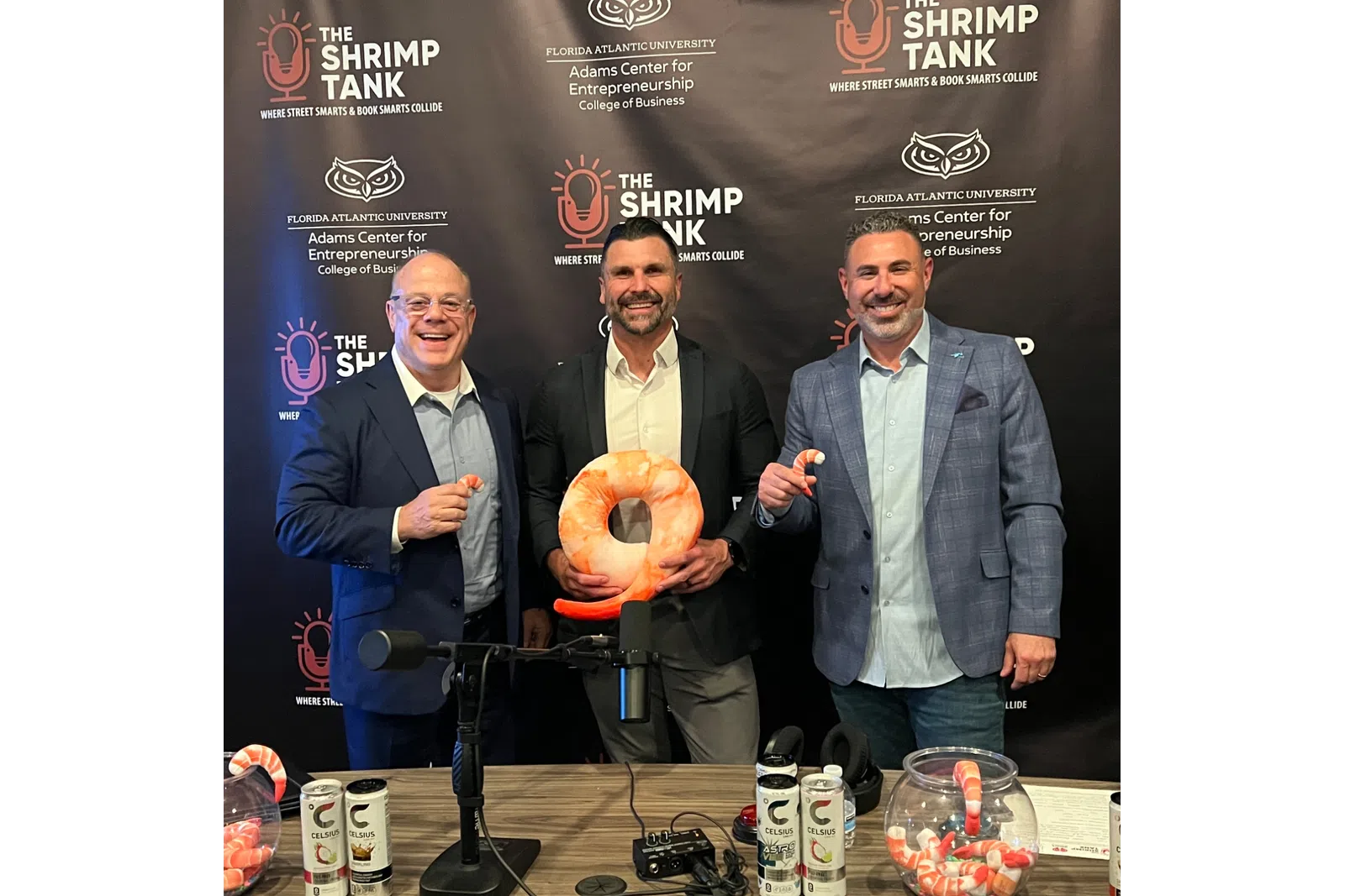 Three men in suits smiling at a Shrimp Tank event with shrimp-shaped props and beverages on a table.
