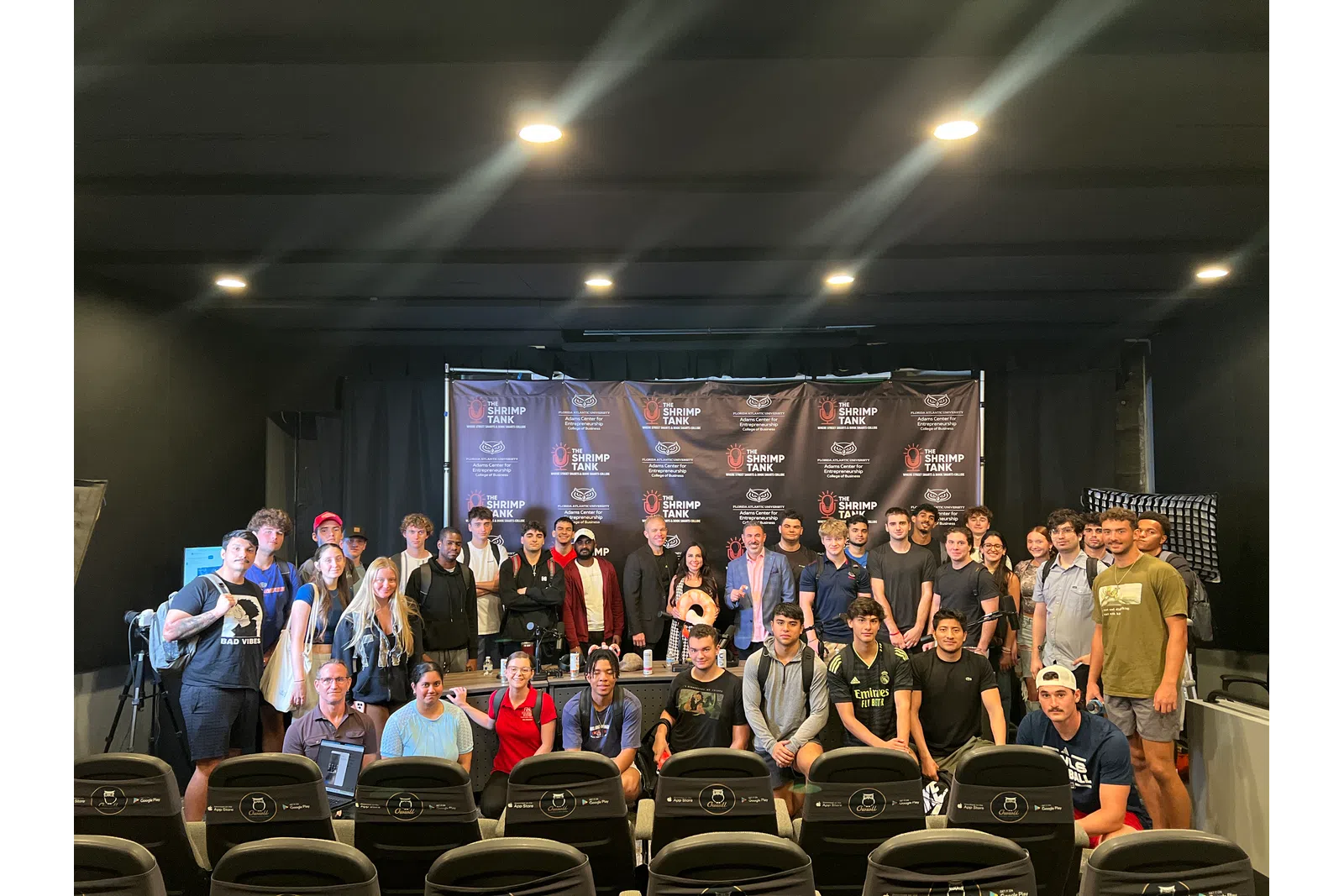 Large group of diverse young adults and professionals posing on stage in front of a Shrimp Tank event banner