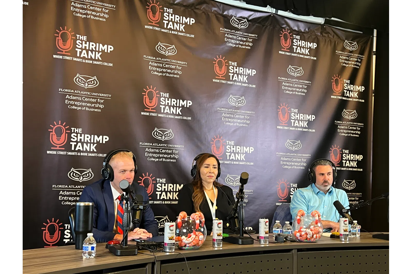 Three hosts speaking into microphones on a podcast set with The Shrimp Tank backdrop and shrimp decor on table.