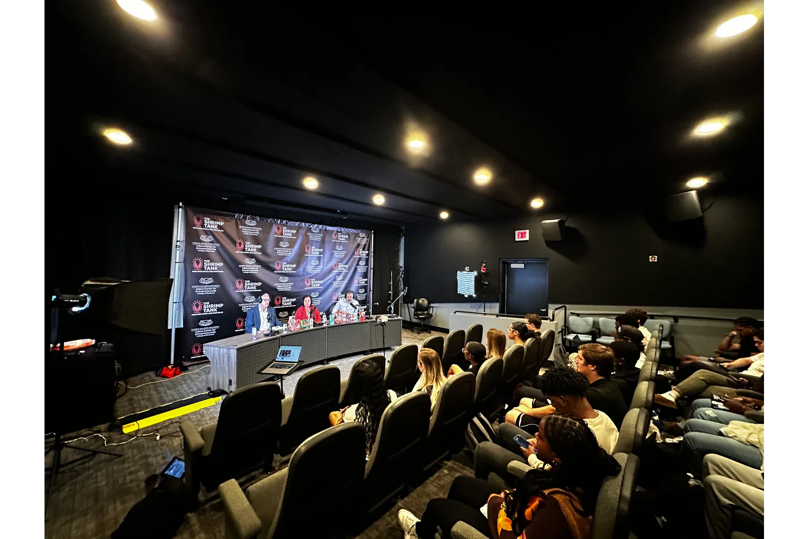 a group of people sitting in a room with a stage and a large screen