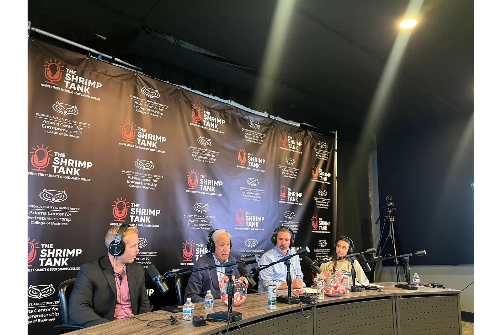Panel of four speakers with microphones in recording studio for The Shrimp Tank podcast with branded backdrop and lighting.