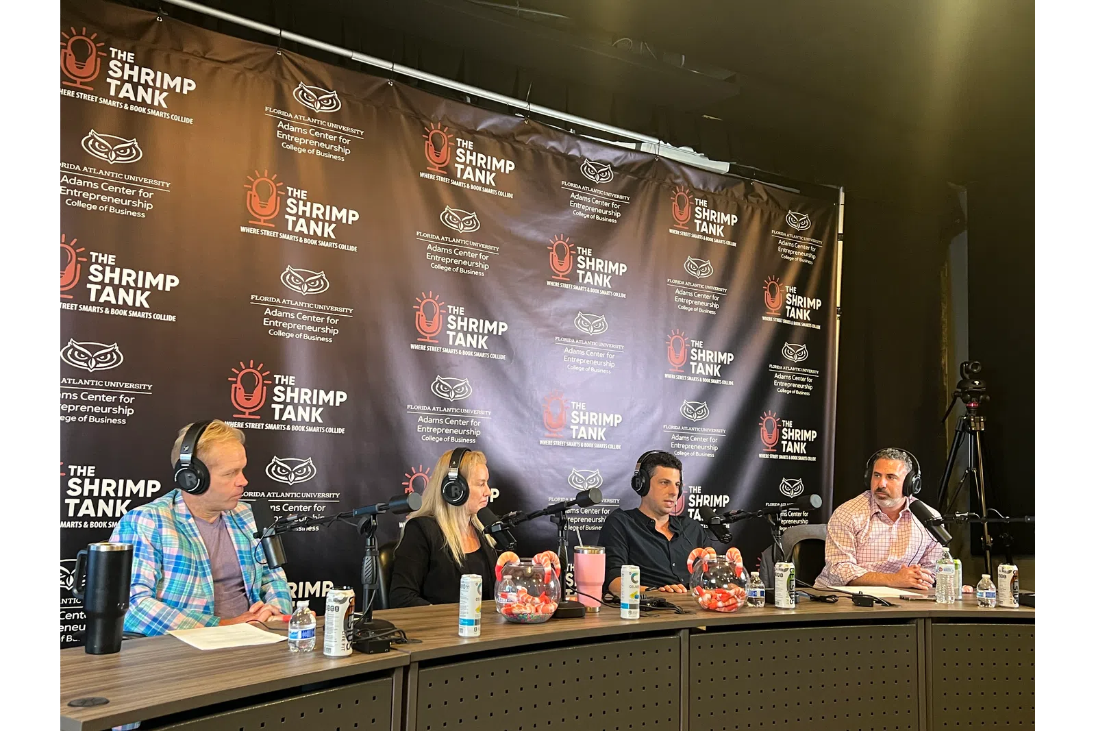 Four hosts wearing headphones speaking into microphones at The Shrimp Tank podcast panel with branded backdrop