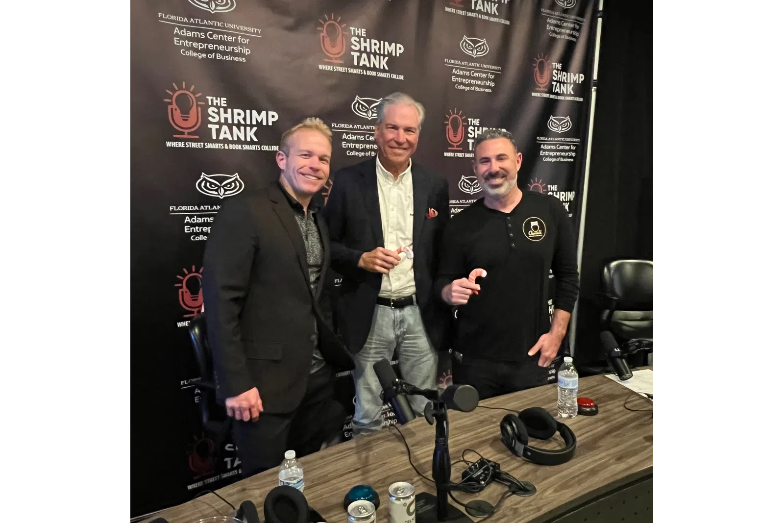 Three men posing in front of The Shrimp Tank podcast backdrop with a table of podcast equipment and drinks.