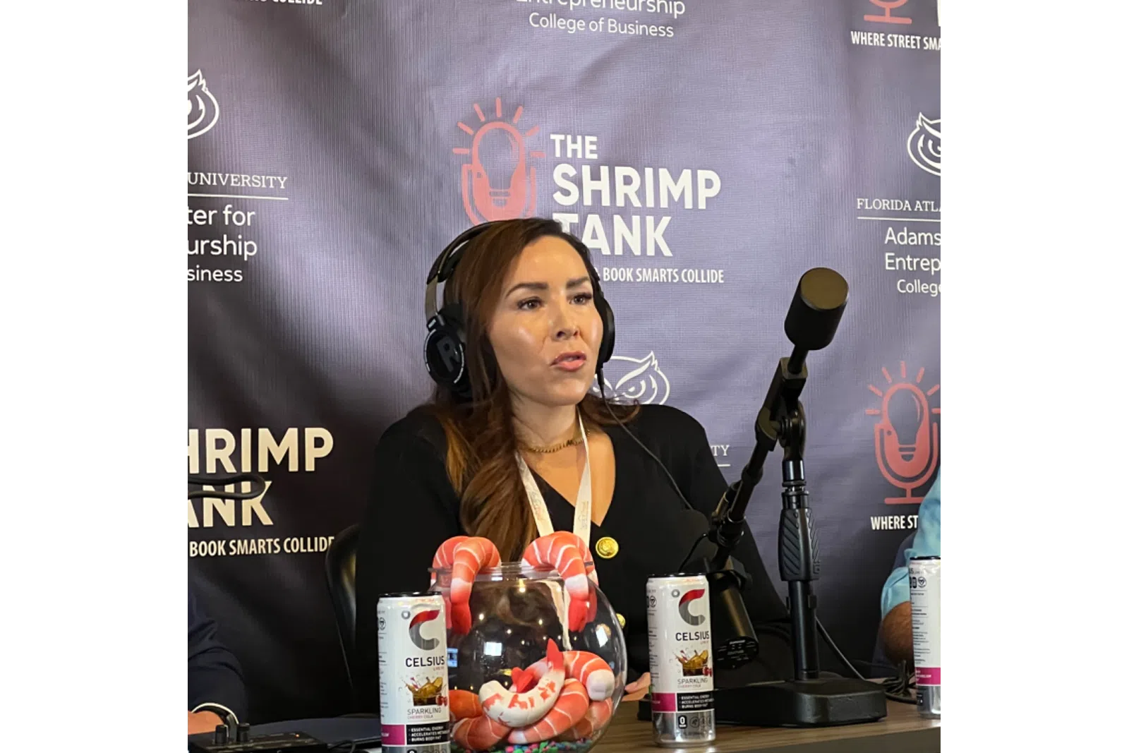 Woman wearing headphones speaks into microphone at The Shrimp Tank event with promotional beverages displayed.