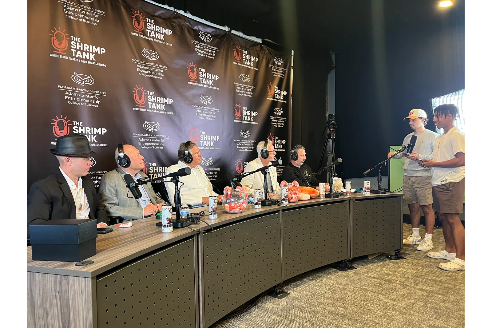 Panel of five men recording a podcast at The Shrimp Tank event with two men hosting on the side.