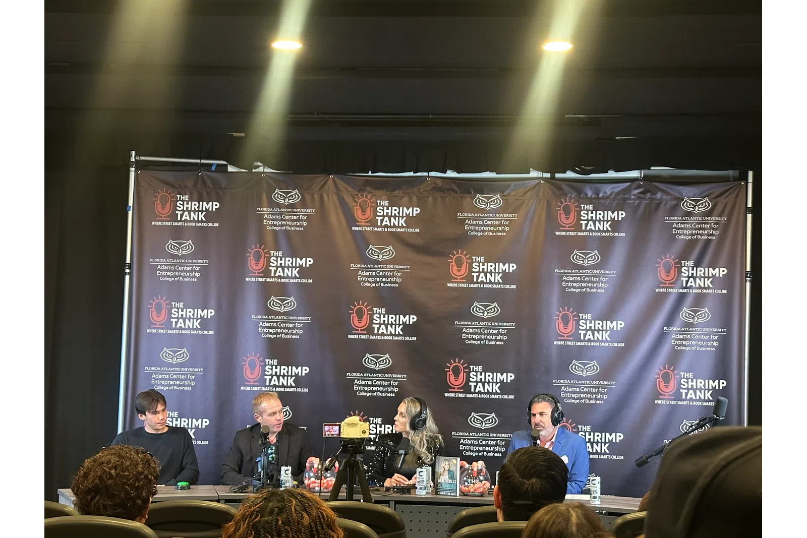Panel of four speakers at The Shrimp Tank event with branded backdrop and audience in foreground