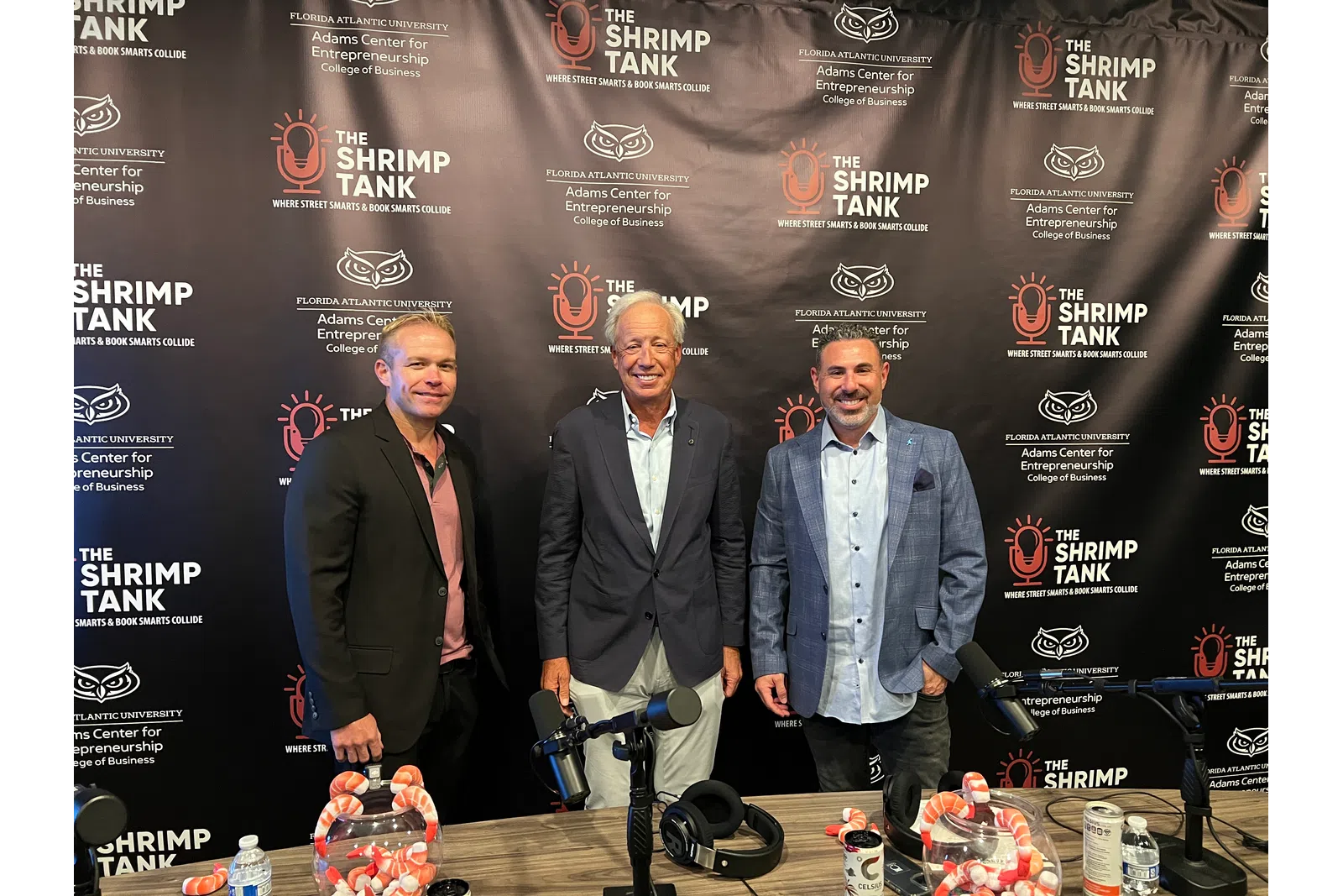 Three men standing in front of The Shrimp Tank podcast backdrop with microphones and headphones on table