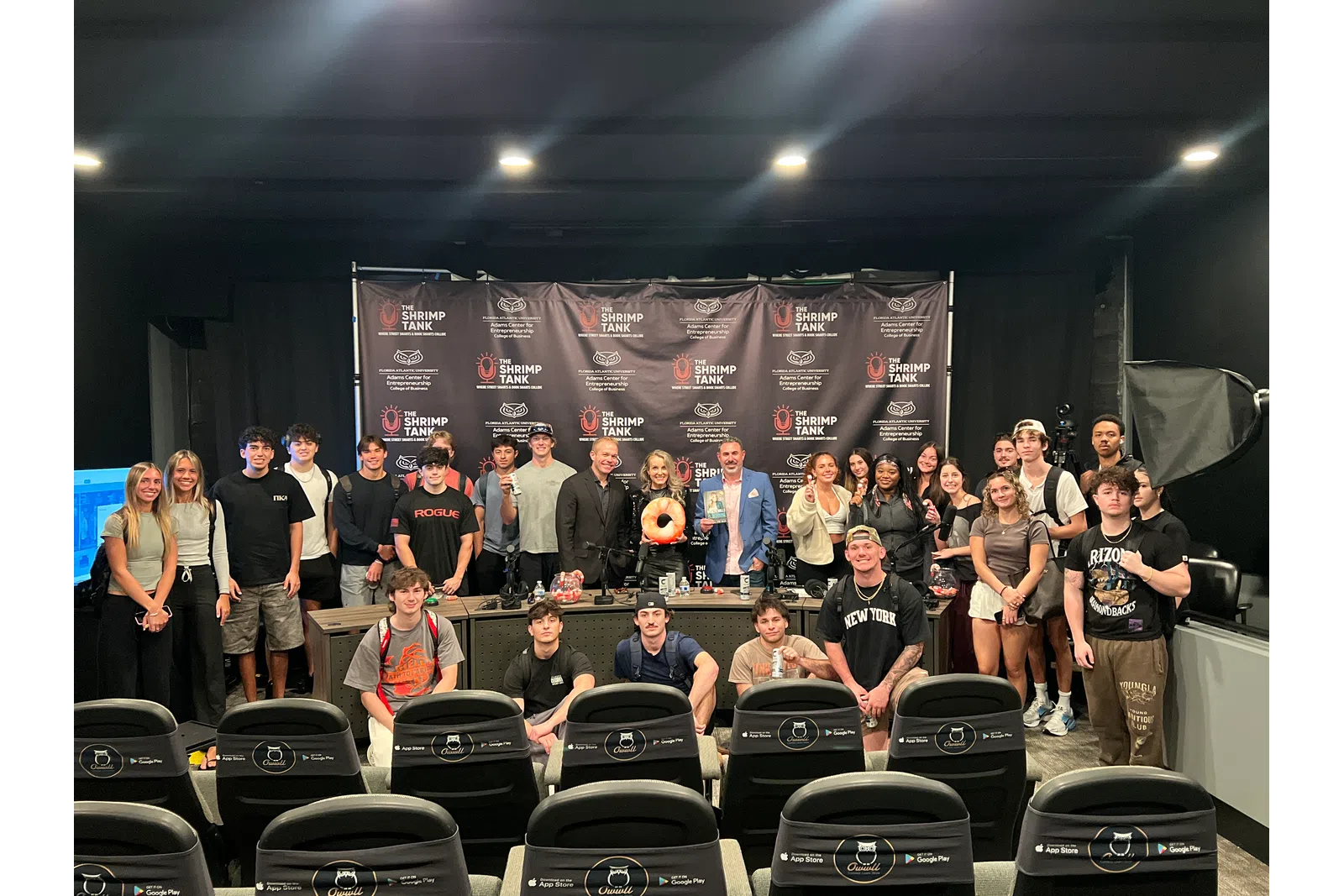Large group of diverse people posing in front of The Shrimp Tank backdrop in a studio setting with chairs in foreground.