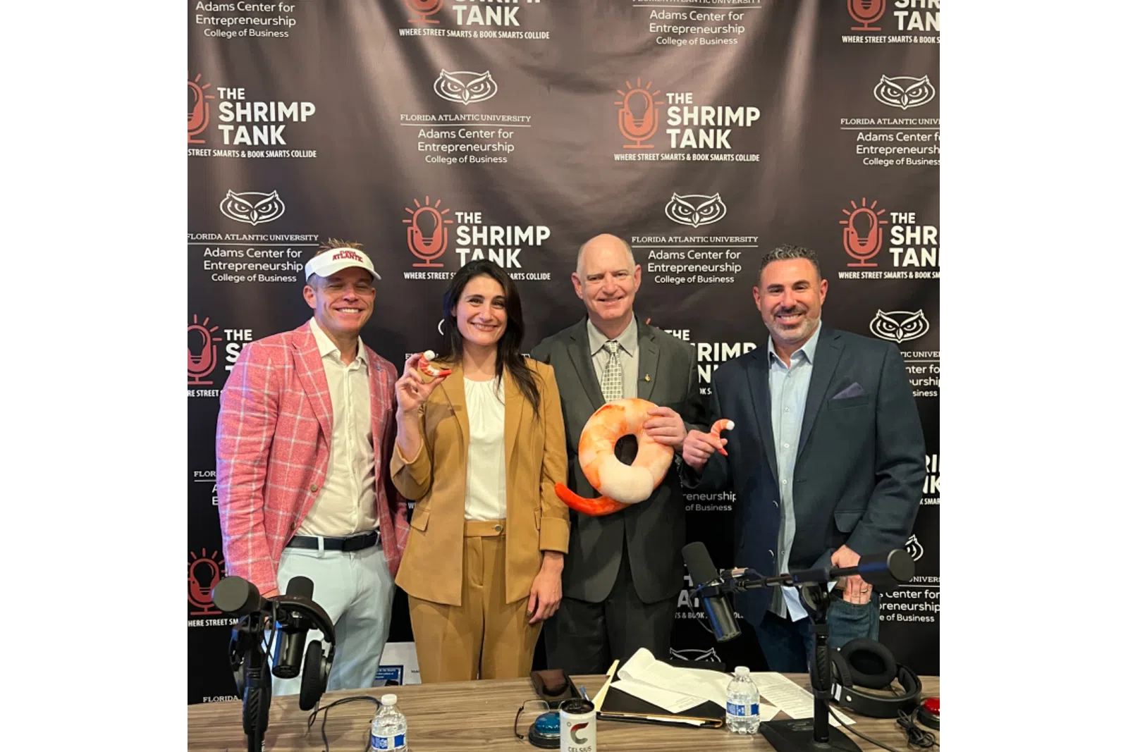 Four people smiling at a shrimp-themed podcast with microphones and branded backdrop for The Shrimp Tank podcast.