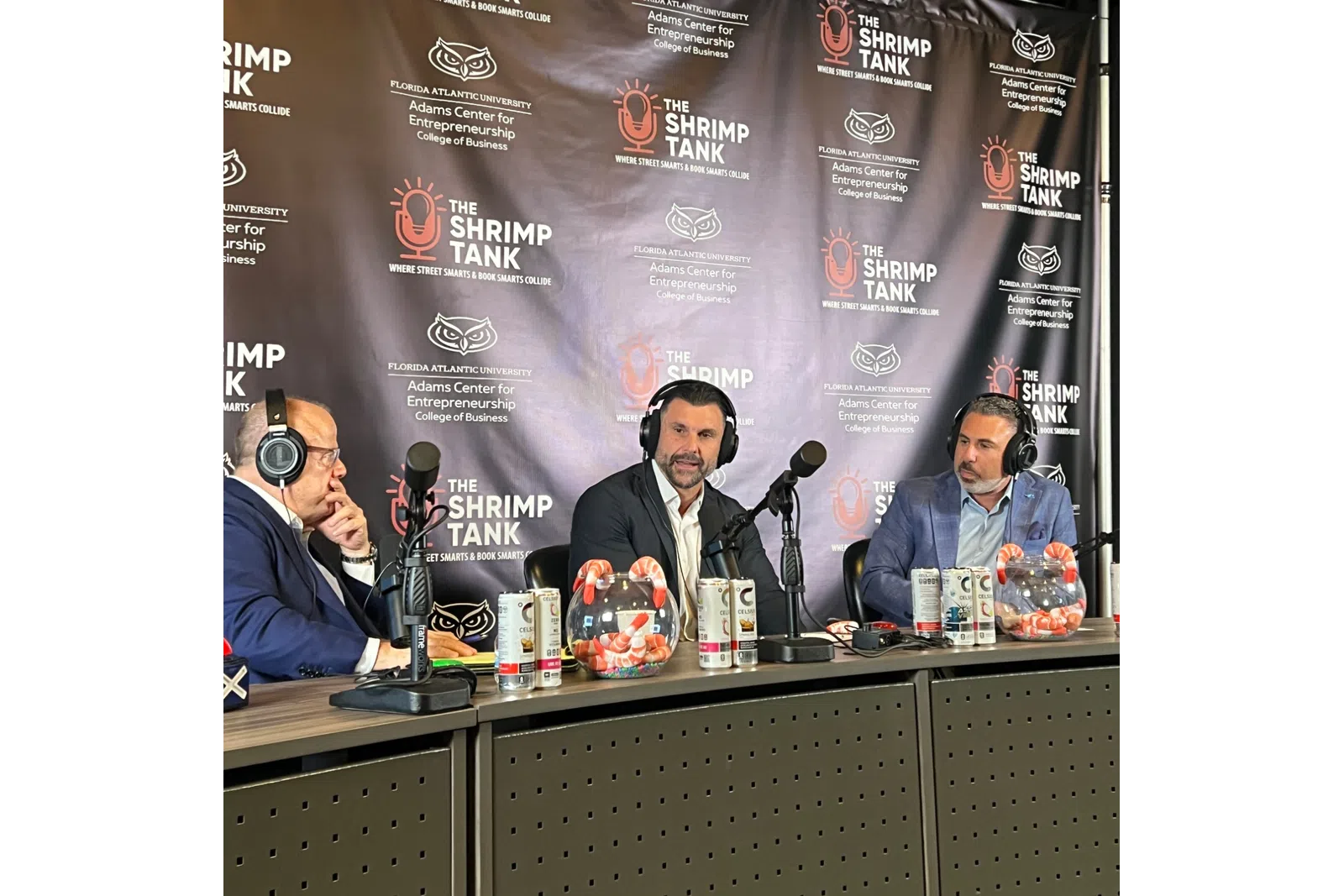Three men wearing headphones sitting at a podcast table with microphones and shrimp tanks beside them.