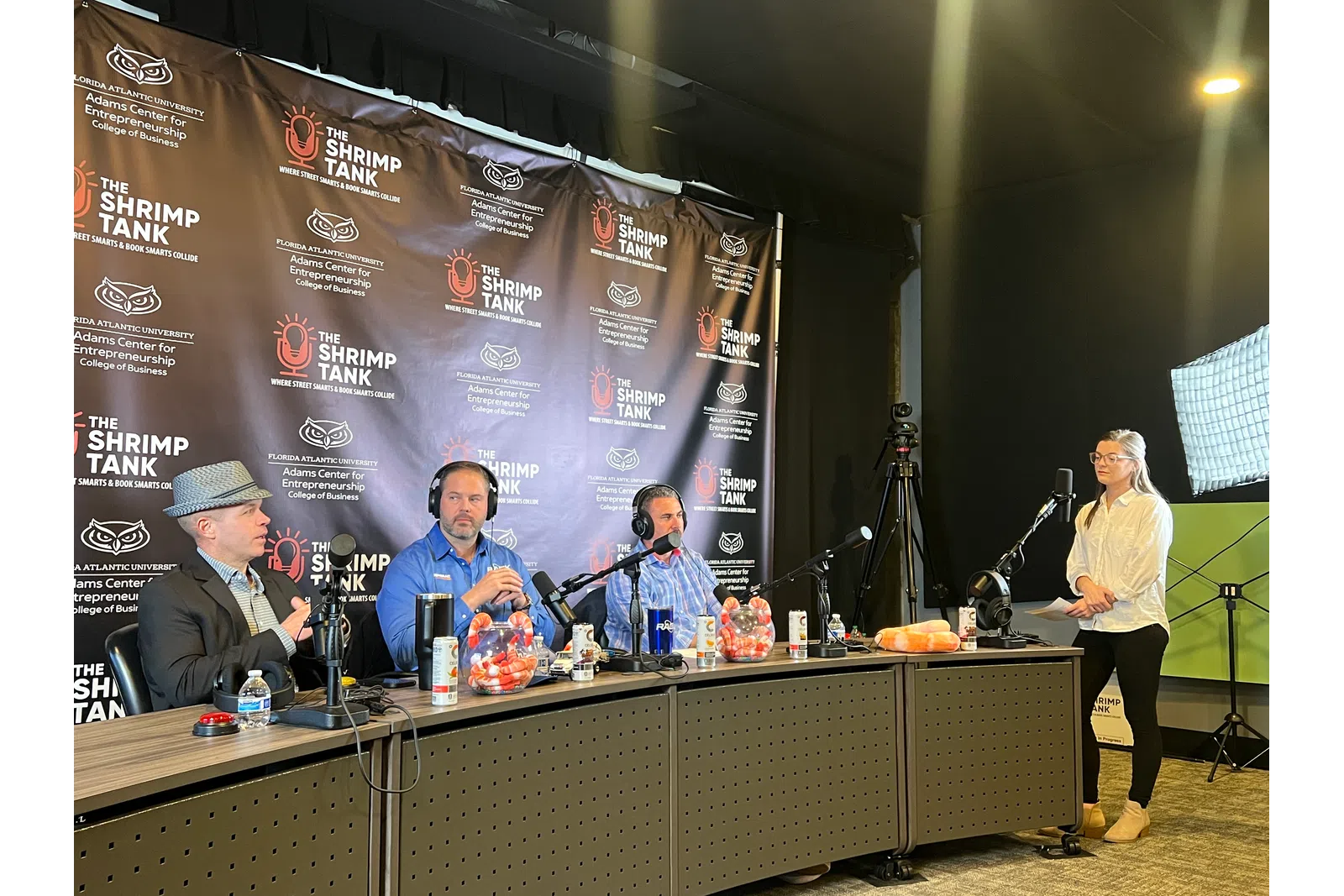 Podcast recording with three men wearing headphones, microphones, and a woman standing by light and camera setup.