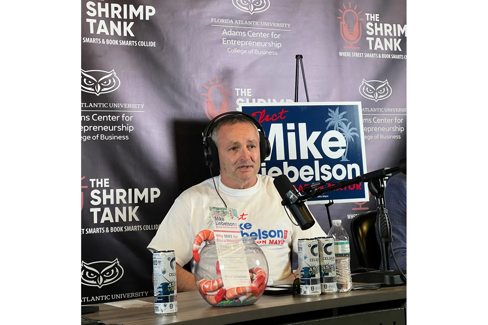 Man wearing headphones speaks into microphone on podcast set with shrimp tank backdrop and campaign sign.