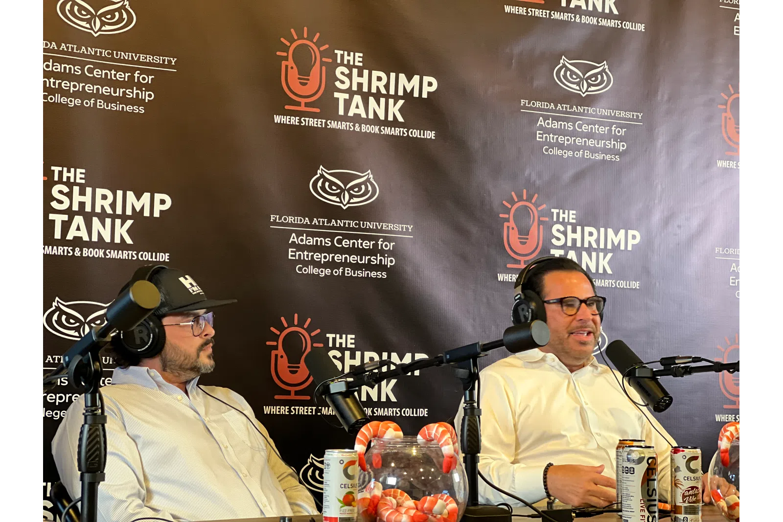 Two men wearing headsets speaking into microphones during a podcast at The Shrimp Tank in front of a branded backdrop.