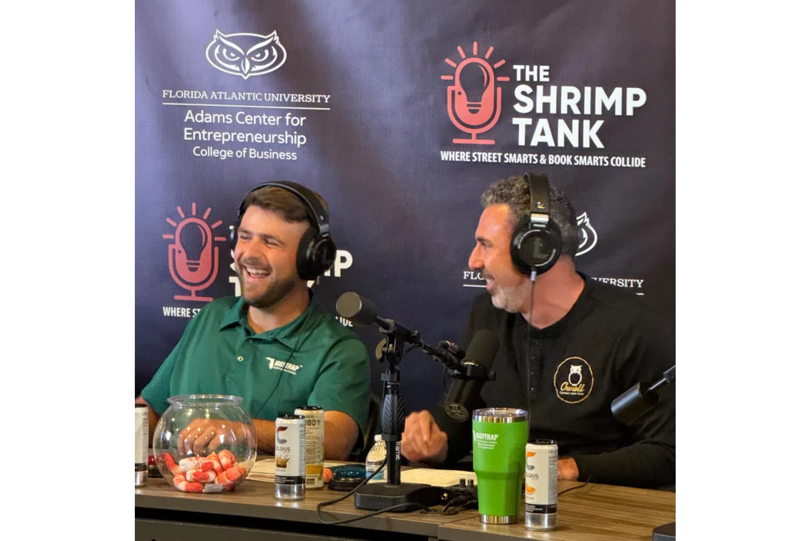 Two men wearing headphones recording a podcast at a table with microphones and drinks in front of a Shrimp Tank backdrop.