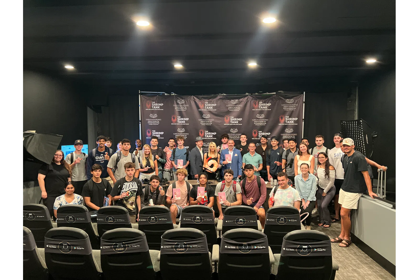 Large group of young adults posing in a small theater with a Shrimp Tank branded backdrop and seats in foreground.