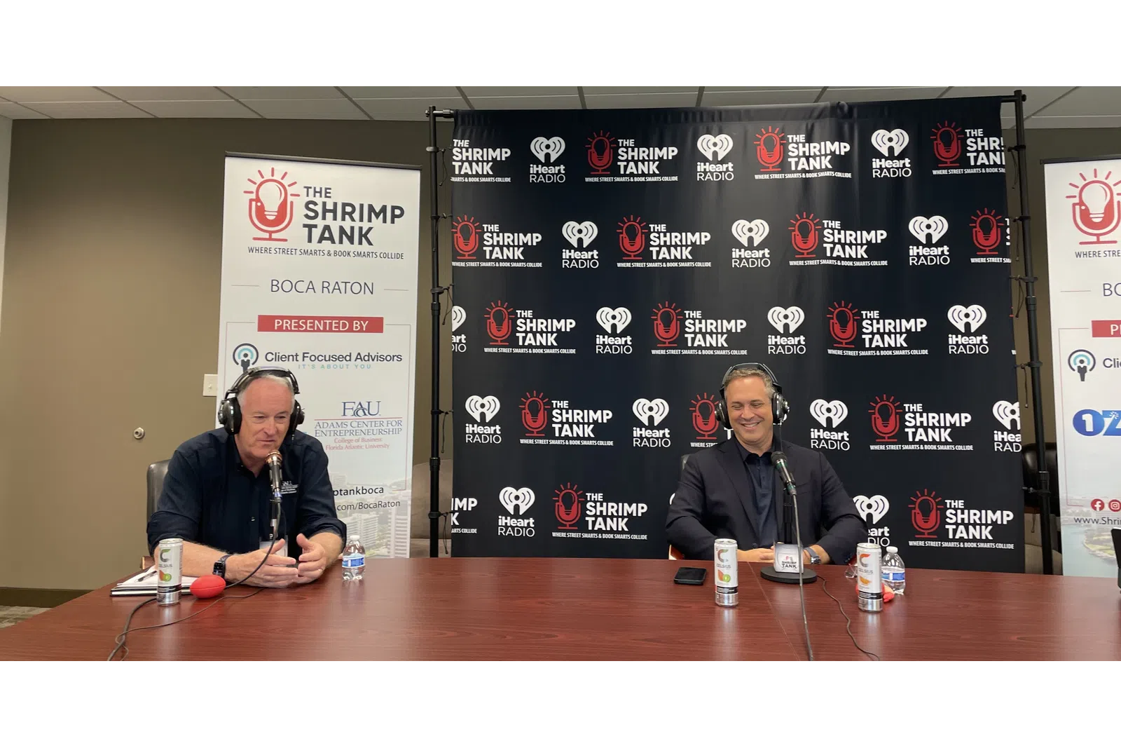 a couple of men sitting at a table with microphones and a sign behind them